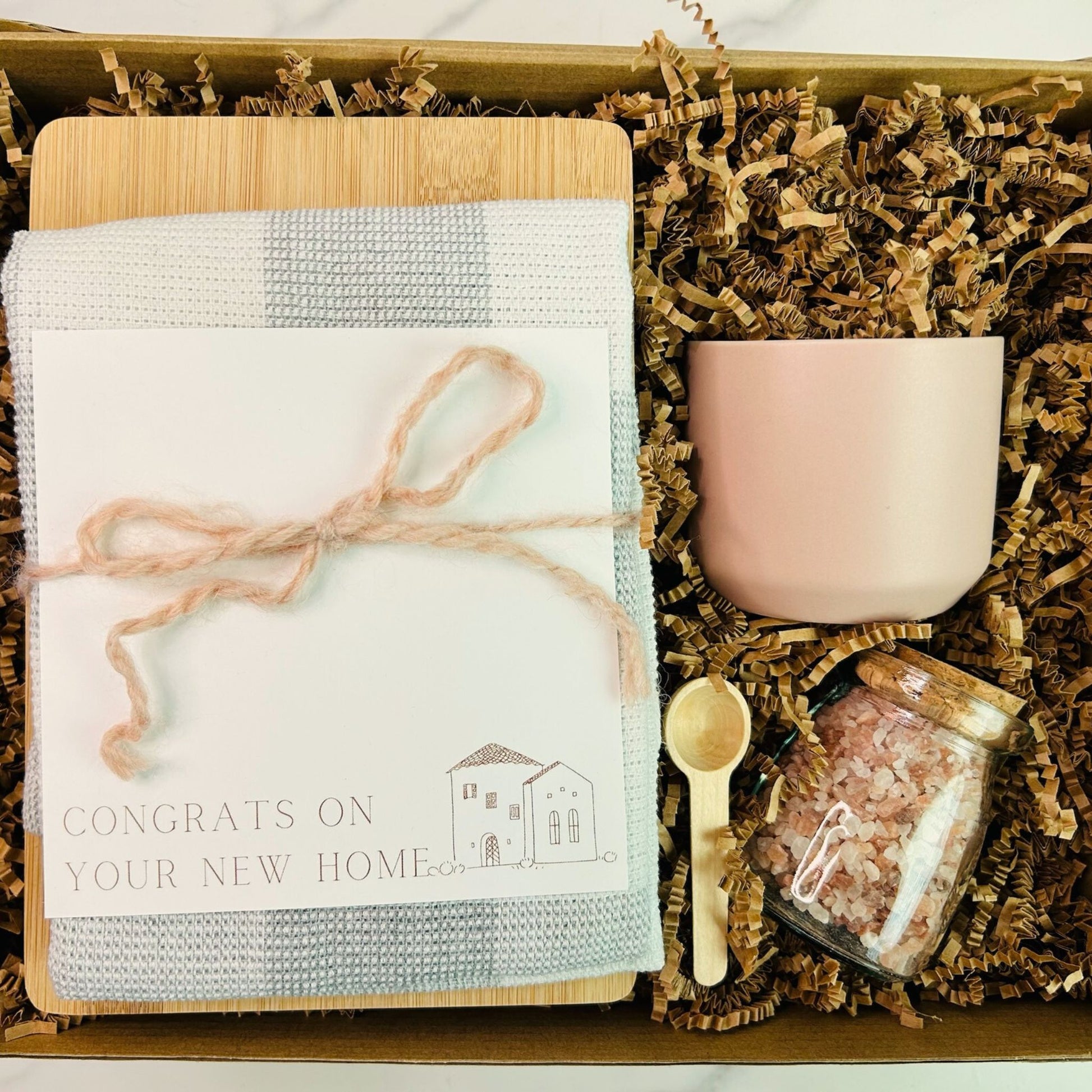 A jar of pink Himalayan salt with a small wooden spoon, a cutting board, and a checkered dish towel, tied together with twine, placed on a kitchen counter. real estate gift, gift for clients, realtor