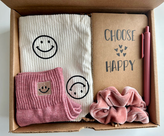 A gift set containing a pair of socks with a smiley face design, a velvet scrunchy, a pen, a notebook with a happy design on the cover, and a smiley face bag.