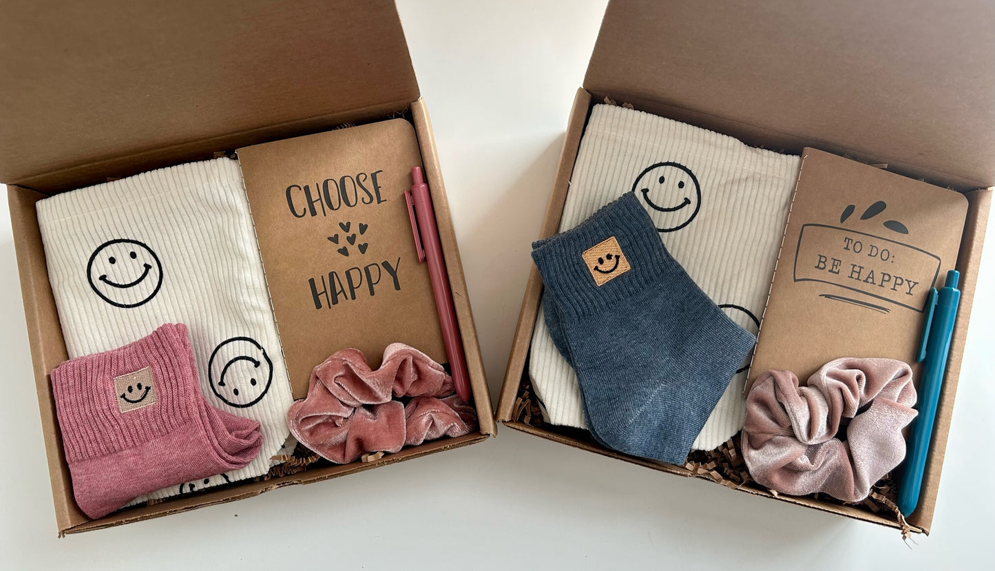 Two options of the Happy Vibes gift box. Includes smiley face bag, happiness journal, pen, scrunchie, and smiley face socks. Colors vary.