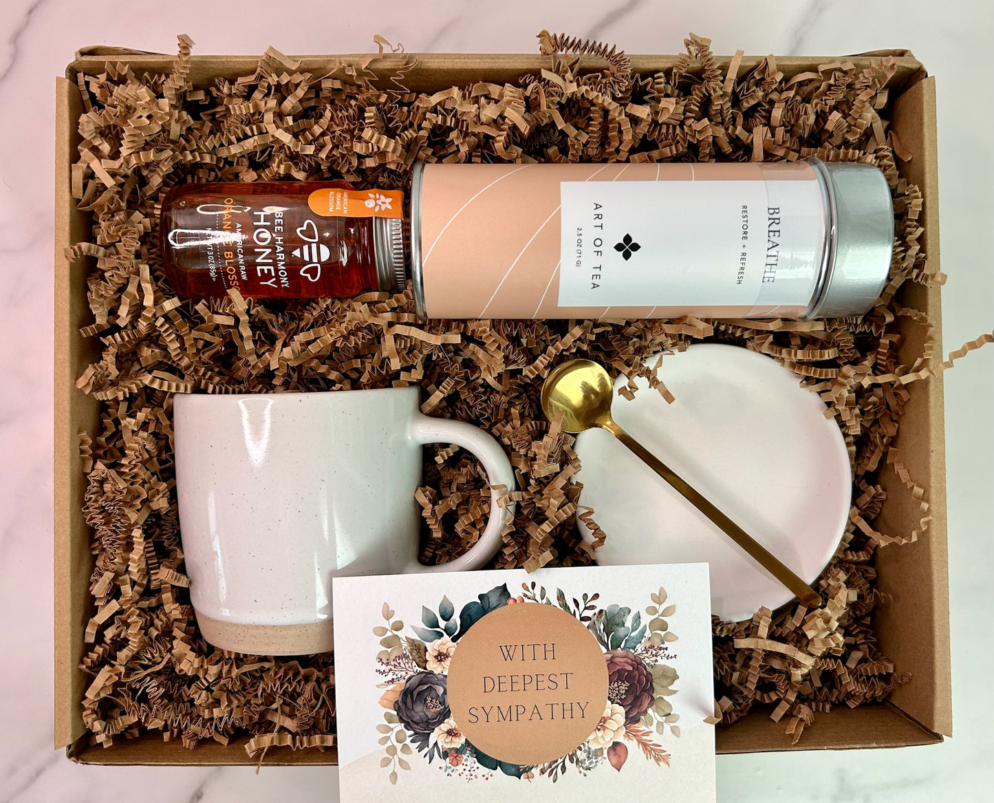 Flat lay of deluxe sympathy gift box including herbal tea, premium honey, speckled mug, gold spoon, ceramic dish, and sympathy card