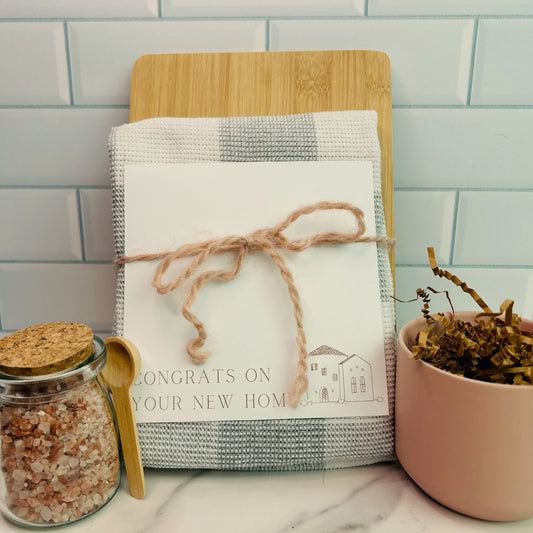 A jar of pink Himalayan salt with a small wooden spoon, a cutting board, and a checkered dish towel, tied together with twine, placed on a kitchen counter. real estate gift, gift for clients, realtor