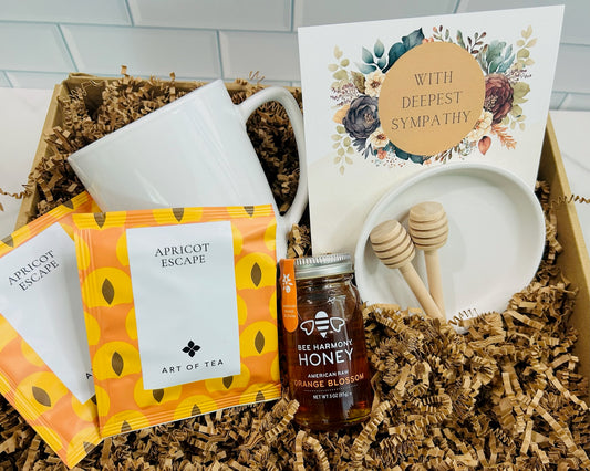A Sympathy Box gift set containing premium raw honey, honey stir sticks, a porcelain dish, and two Art of Tea Apricot Escape satchels, presented in an embellished kraft box with a card expressing sympathy.
