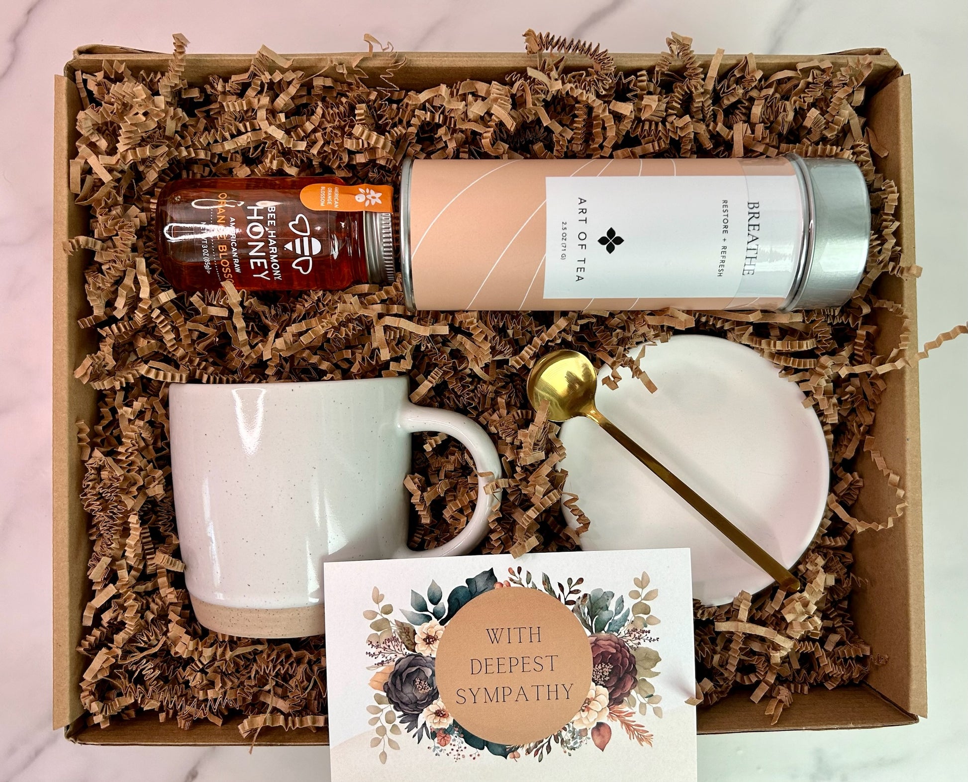Flat lay of deluxe sympathy gift box including herbal tea, premium honey, speckled mug, gold spoon, ceramic dish, and sympathy card