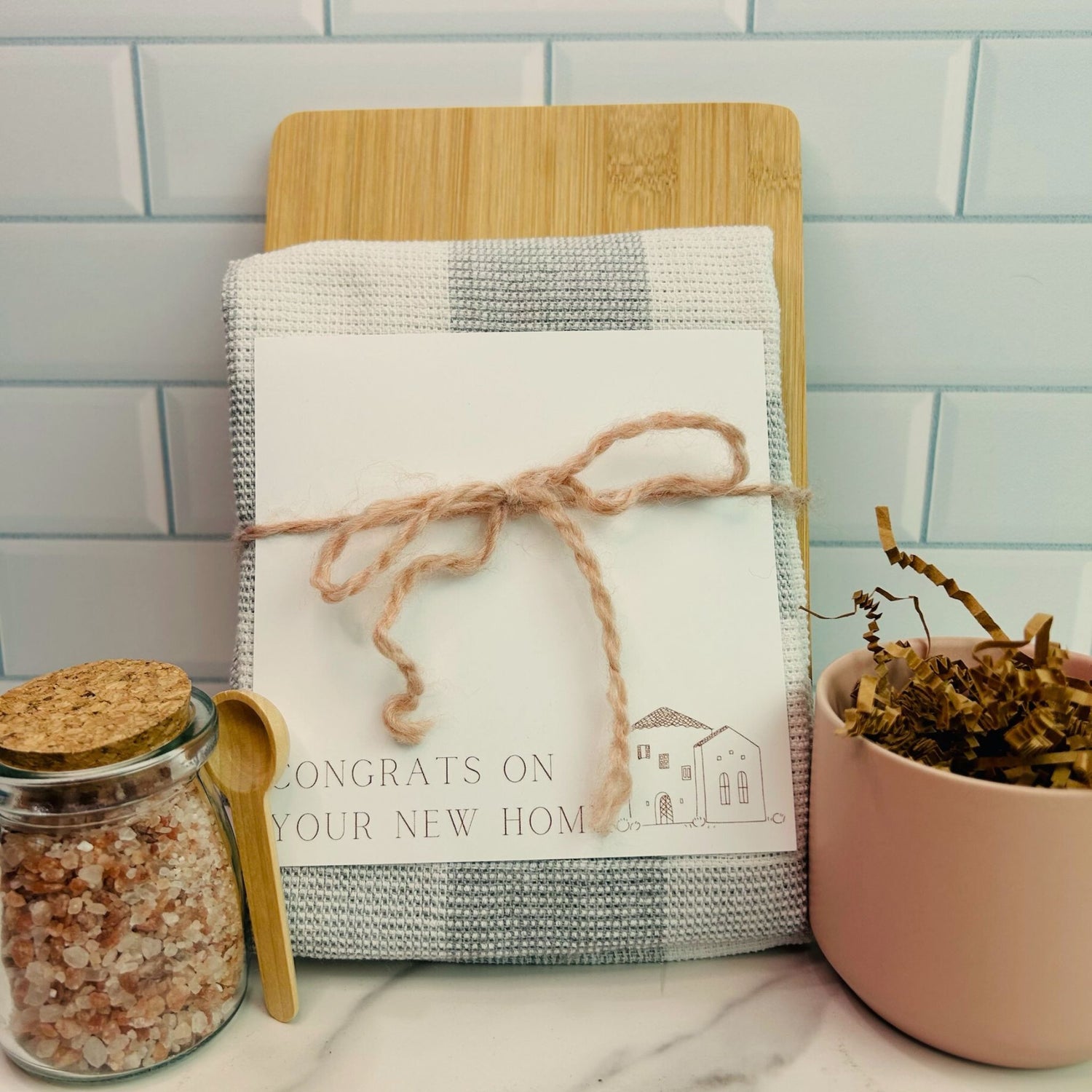 A jar of pink Himalayan salt with a small wooden spoon, a cutting board, and a checkered dish towel, tied together with twine, placed on a kitchen counter. real estate gift, gift for clients, realtor