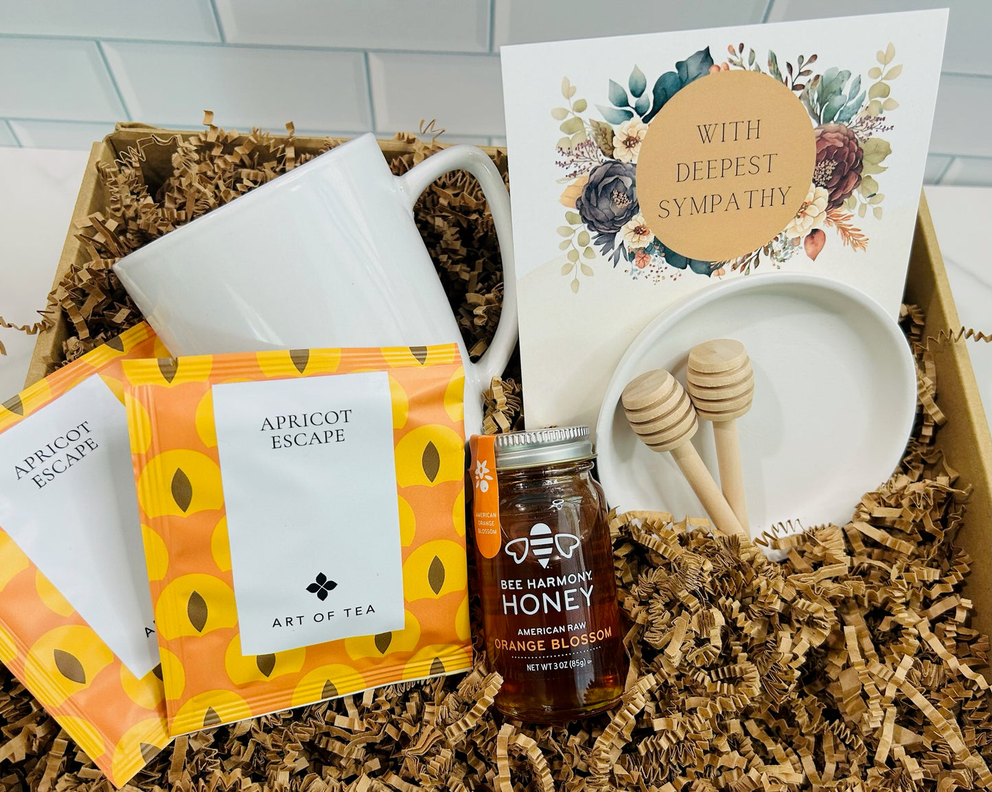 A Sympathy Box gift set containing premium raw honey, honey stir sticks, a porcelain dish, and two Art of Tea Apricot Escape satchels, presented in an embellished kraft box with a card expressing sympathy.